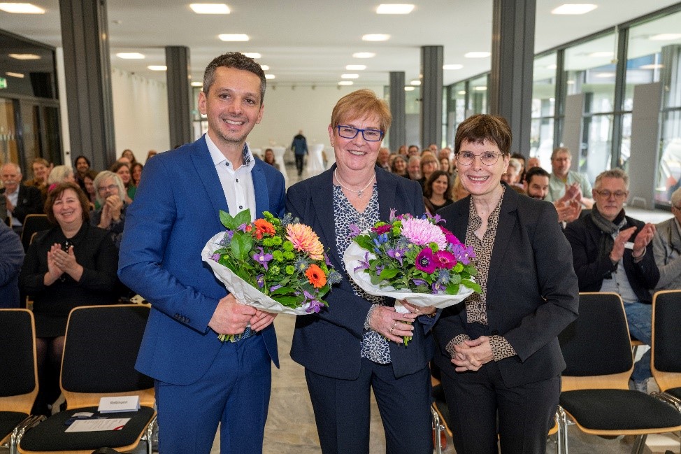 Drei Personen stehen in der vorderen Reihe eines Veranstaltungsraums und halten jeweils einen Blumenstrauß in den Händen. Sie lächeln in die Kamera. Links im Bild steht ein Mann im blauen Anzug, daneben zwei Frauen in dunkler Kleidung. Hinter ihnen sitzen zahlreiche Menschen im Publikum, die applaudieren.
