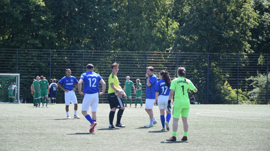 Fußballspieler in blauen und grünen Trikots auf einem Spielfeld mit Tor und Bäumen im Hintergrund