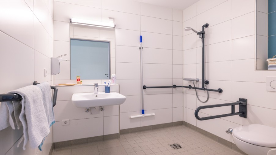 Ein mit Hilfsmitteln ausgestattetes Badezimmer mit ebenerdiger Dusche und unterfahrbarem Waschbecken.