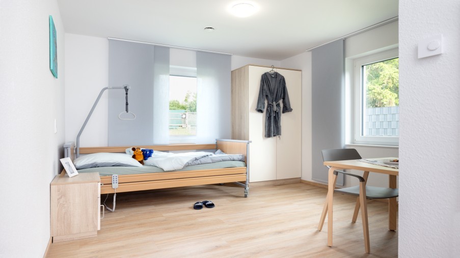 Ein licht durchflutetes voll möbliertes Zimmer mit Pflegebett, Nachttisch, Schrank, Tisch und Stuhl sind zu sehen.