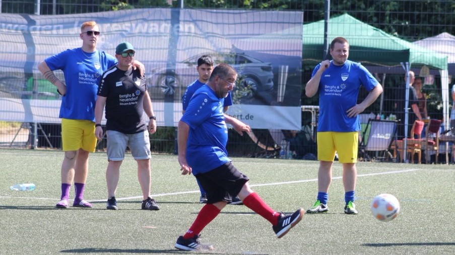 Fußballspieler in Blau und Gelb. Einer schießt den Ball, im Hintergrund stehen Zuschauer und Bäume.