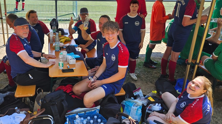 Gruppe von Fußballspielern in blauen Trikots sitzt und steht unter einem Pavillon neben einem Fußballfeld