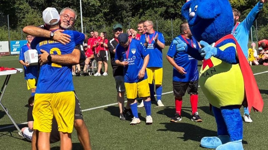 Zwei Spieler in Gelb-Blau umarmen sich. Im Hintergrund stehen weitere Spieler und ein blaues Maskottchen.
