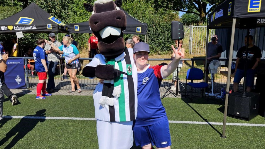 Person in blau-rotem Sportoutfit posiert mit einem Maskottchen im schwarz-weiß-grünen Trikot auf dem Rasen.
