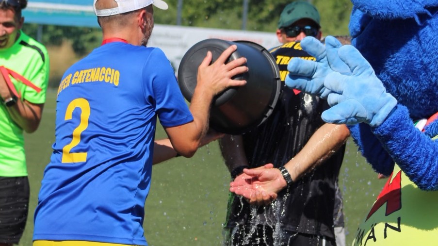 Mann im blauen Trikot schüttet Wasser über einen Mann in Schwarz; daneben steht ein blaues Maskottchen.

