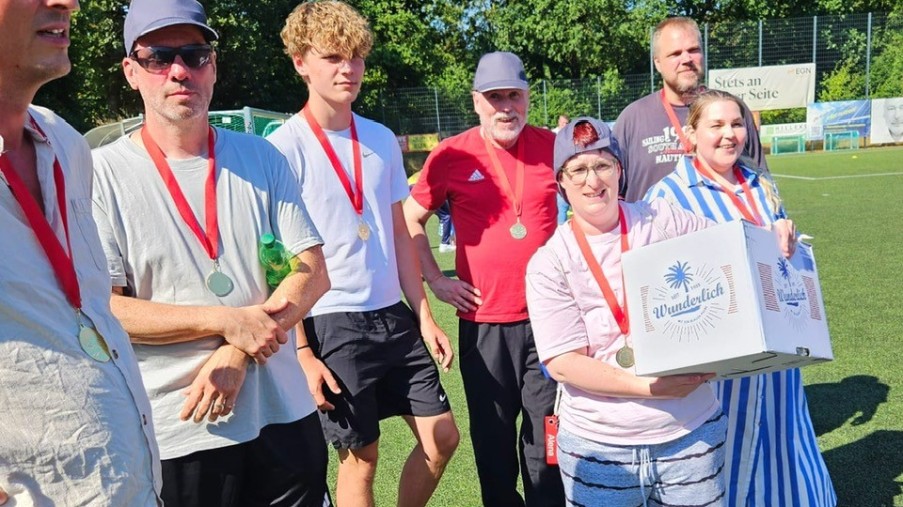 Gruppe von Menschen mit Medaillen steht auf einem Sportplatz, eine Frau hält einen Karton mit Aufdruck