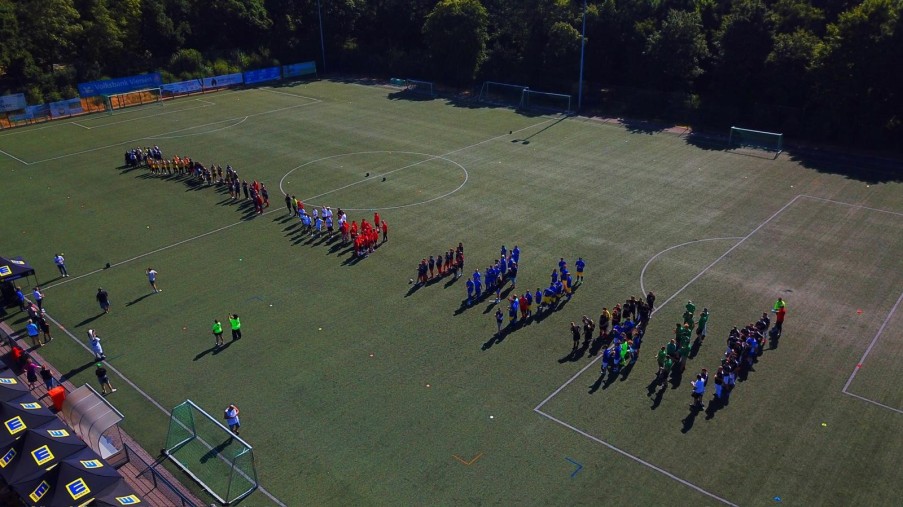Mehrere Fußballmannschaften in unterschiedlichen Trikots stehen in Reihen auf einem Spielfeld im Freien
