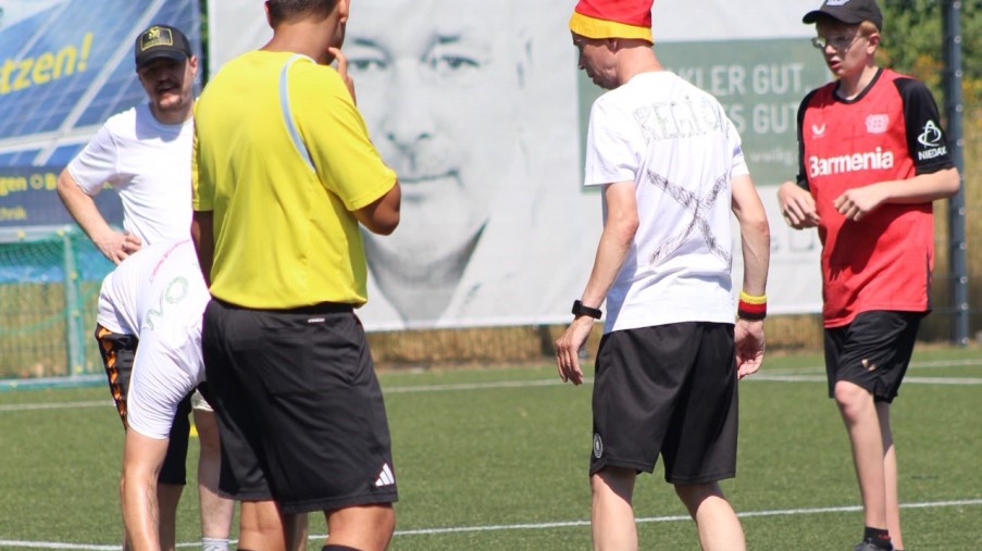 Mehrere Männer auf einem Fußballfeld. Einer trägt ein gelbes Trikot, eine Person beugt sich zum Ball.
