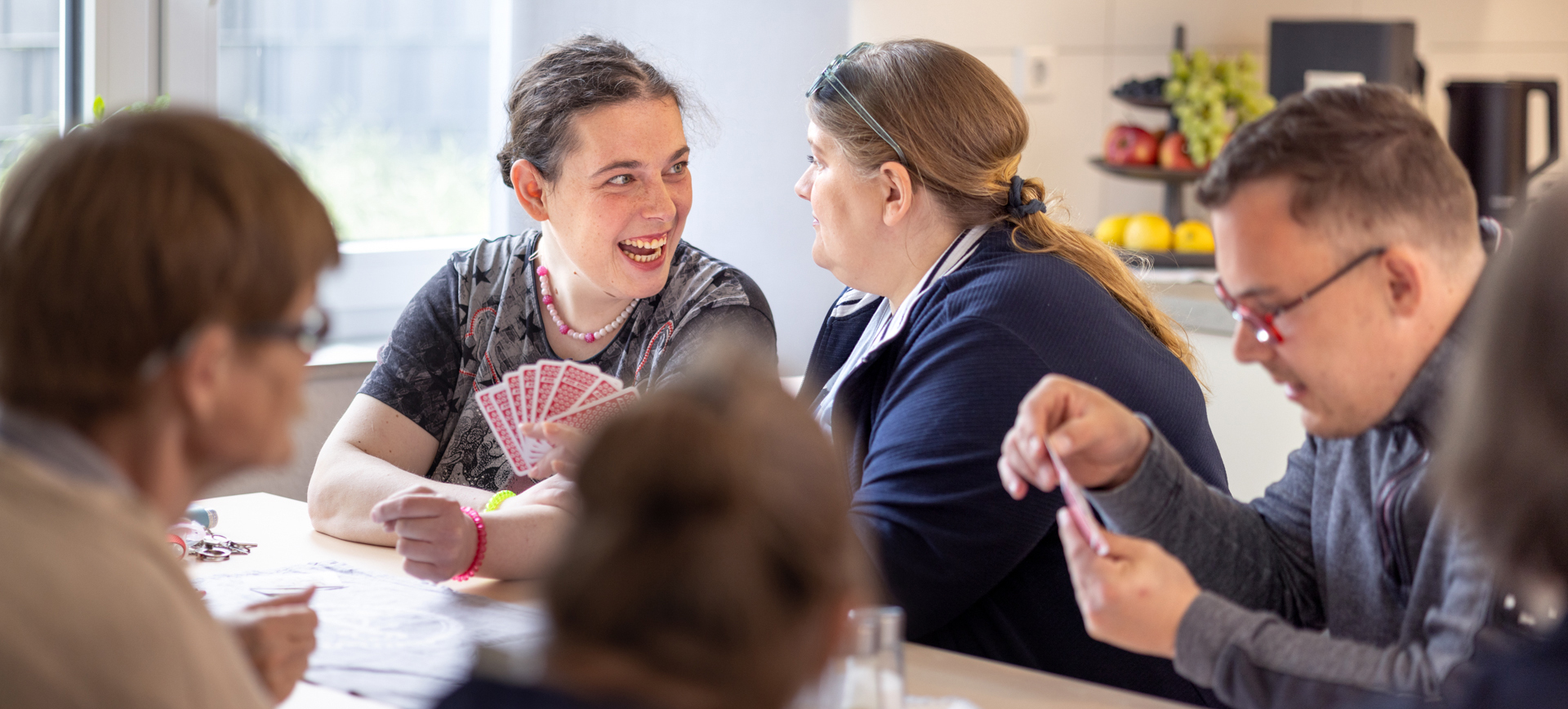 Mehrere Menschen, die in einer besonderen Wohnform leben, sitzen um einen Tisch herum und spielen gemeinschaftlich Karten.