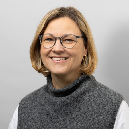 Eine Frau mit hellbraunen Haaren und einer grauen Brille lächelt mich sichtbaren Zähnen in die Kamera.