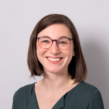 Eine Frau mit dunklen schulterlangen Haaren und einer rundlichen Brille lächelt mich sichtbaren Zähnen in die Kamera.